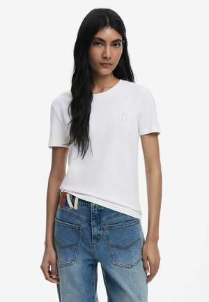 T-shirt en coton blanc avec un col rond, des manches courtes et un petit détail brodé en forme de 'D' sur la poitrine gauche. Porté avec un jean en denim bleu.