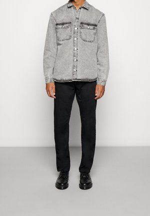 Homme portant une veste en jean gris clair boutonnée, un pantalon noir et des chaussures à lacets noires, debout devant un fond clair uni.