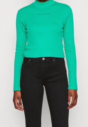 Personne portant un haut à manches longues vert vif avec le texte "MARC O'POLO DENIM" et un jean noir taille haute sur fond blanc.