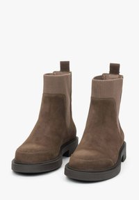 Para brązowych botków zamszowych z elastycznymi panelami bocznymi i pętelkami do zakładania, na czarnych gumowych podeszwach, przedstawionych na białym tle.