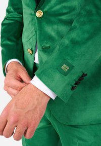 Žalias aksominis švarkas su auksinėmis sagomis, juodos sagos detalės ant rankogalių ir etiketė su užrašu "OPPO SUITS" ant rankovės.