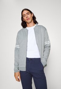 Chaqueta bomber gris con rayas blancas en las mangas, cierre frontal, y puños de canalé, llevada sobre una camiseta blanca y pantalones navy.