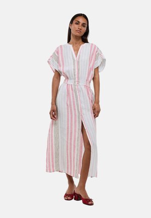 Femme portant une robe blanche avec des rayures verticales roses et noires et une fente à l'avant, associée à des sandales rouges à talons, debout sur un fond uni.
