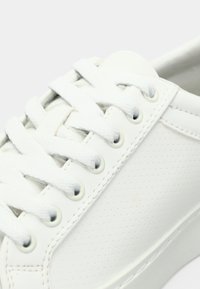 Valge nahast spordijalats tekstuuriga pinnaga, lame valge paeltega ja silmadega. Sile toe-kate ja pehmendatud krae. Perforeeritud külgpaneeli detail.