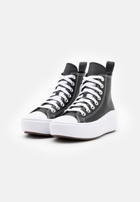 Svarte høyhælte sneakers laget av teksturert materiale, med hvite snører, en tykk hvit såle og en kontrastfarget gummitåhette.