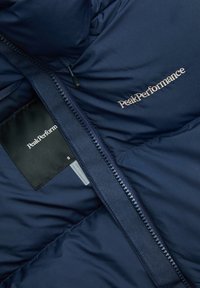 Marineblaue Steppjacke mit einem quiltierten Innenfutter, ausgestattet mit einem Reißverschluss und gesticktem "PeakPerformance"-Logo. Pflegeetikett sichtbar.