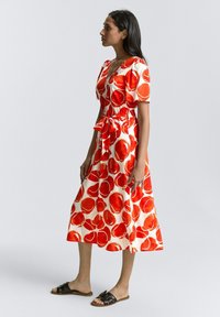 Robe imprimée rouge et blanche avec un design croisé, manches courtes et une ceinture assortie, associée à des sandales à glissière noires en tissu lisse.