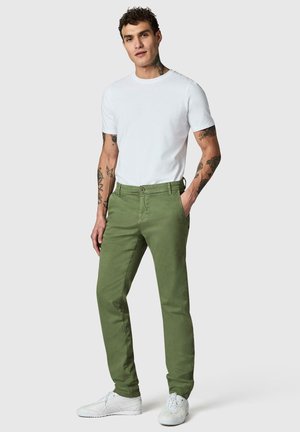 Junger Mann mit Tattoos, der ein weißes kurzärmeliges Hemd, olivgrüne Hose und weiße Sneakers trägt und mit einer Hand in der Tasche steht.