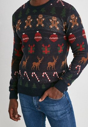 Maglione natalizio da uomo blu scuro con omini di pan di zenzero, decorazioni, regali, renne, bastoncini di zucchero e alberi di Natale, indossato con jeans blu.