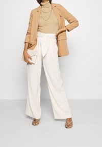 Blazer beige sur un haut côtelé beige, associé à un pantalon blanc à jambes larges noué à la taille et des sandales ornées. Pochette à la main.