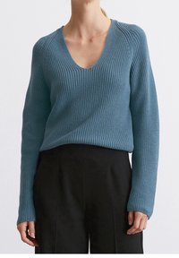Femme portant un pull bleu en maille côtelée avec col en V et un pantalon noir, debout devant un fond clair uni.