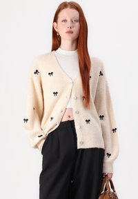 Gilet en crème avec broderie de nœud noir, en tissu tricoté texturé, col en V, coupe oversize, cinq boutons et épaules tombantes, associé à un pantalon noir.