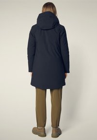 Marine parka met een capuchon, met een lange rechte snit. Gecombineerd met losse olijfgroene broeken en dikke bruine laarzen. Soepele textuur.