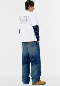 Witte katoenen t-shirt met een afbeelding op de achterkant, gedragen over een marineblauwe gestreepte lange mouwen shirt. Losse blauwe denim jeans en tan schoenen.