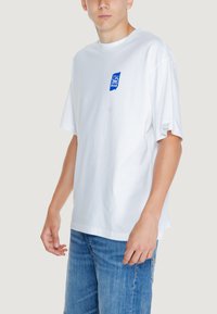 Camiseta de algodón blanca con mangas cortas, que presenta un parche de logo azul en el lado izquierdo del pecho y un corte relajado. Diseñada con un escote redondo.