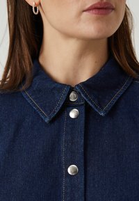 Temno denim srajca z klasičnim ovratnikom, srebrnimi gumbi in kontrastnim šivom, z gladko teksturo in brez vzorcev.