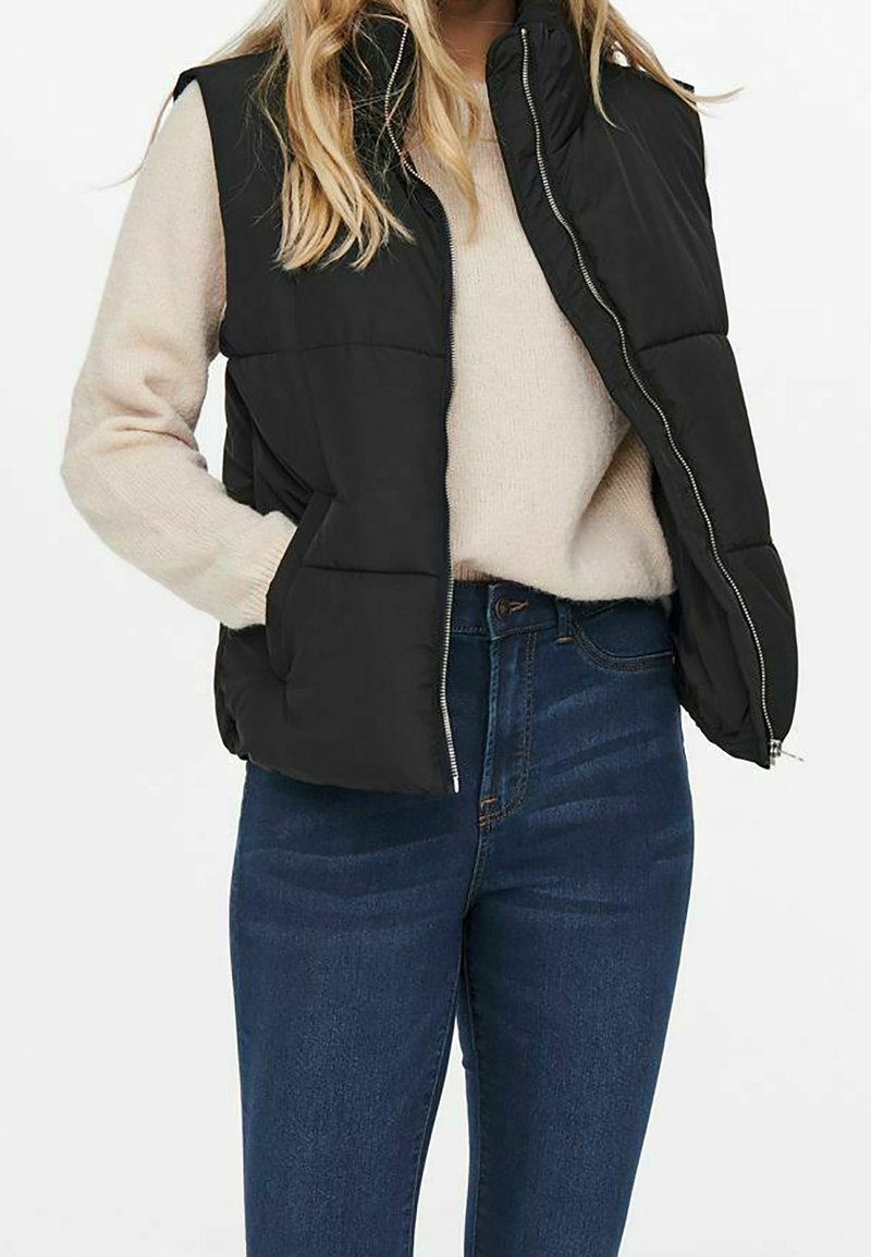 Gilet imbottito nero con cerniera frontale, collo alto e tasche laterali, abbinato a un maglione chiaro e jeans blu scuro. Texture liscia, design casual.