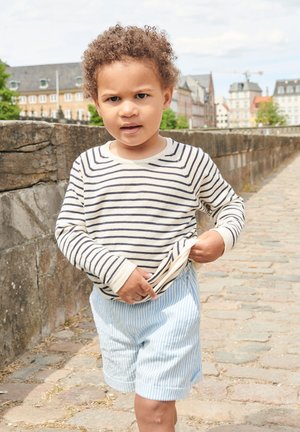 Junges Kind mit lockigem Haar, das ein gestreiftes Langarmshirt und Shorts trägt, läuft auf einem Kopfsteinpflasterweg nahe einer Steinmauer in einer Stadt.