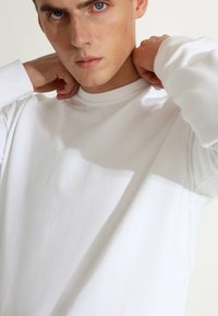 Bjela košulja dugih rukava izrađena od teksturirane tkanine, sa okruglim izrezom, rebrastim manžetama i opuštenim krojem. Vidljive sjene na tkanini.