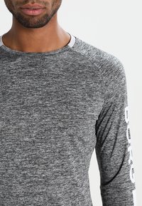 Man wearing a gray long-sleeve shirt with white text on the sleeve, shown from shoulders to mid-torso against a plain background.