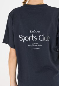 T-paita tummansinisestä puuvillasta, jossa on lyhyet hihat ja valkoisella painettuna teksti: "Les' Arcs Sports Club Luxury Athleisure Wear" selässä.
