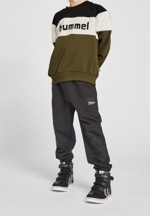 Felpa a strisce verde scuro, nera e bianca con testo "Hummel"; abbinata a pantaloni sportivi neri con toppa del logo e sneakers alte nere.