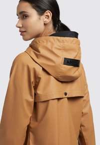 Braune wasserdichte Jacke mit Kapuze, ausgestattet mit einem schwarzen Logo-Tag, schlankem Design und glatter Textur. Knöpfe auf der Rückseite für Verstellbarkeit.