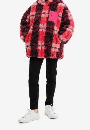 Czerwona i różowa kurtka polarowa w kratę, z zamkiem błyskawicznym i różową kieszenią na piersi. Połączona z czarnymi spodniami i białymi trampkami.