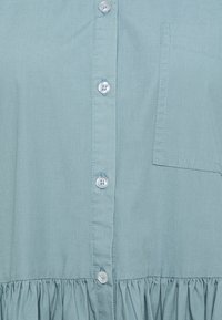 Camisa de botones azul claro con tejido texturizado, que presenta un bolsillo de parche y un dobladillo con volantes. Los botones son transparentes con un ligero brillo.
