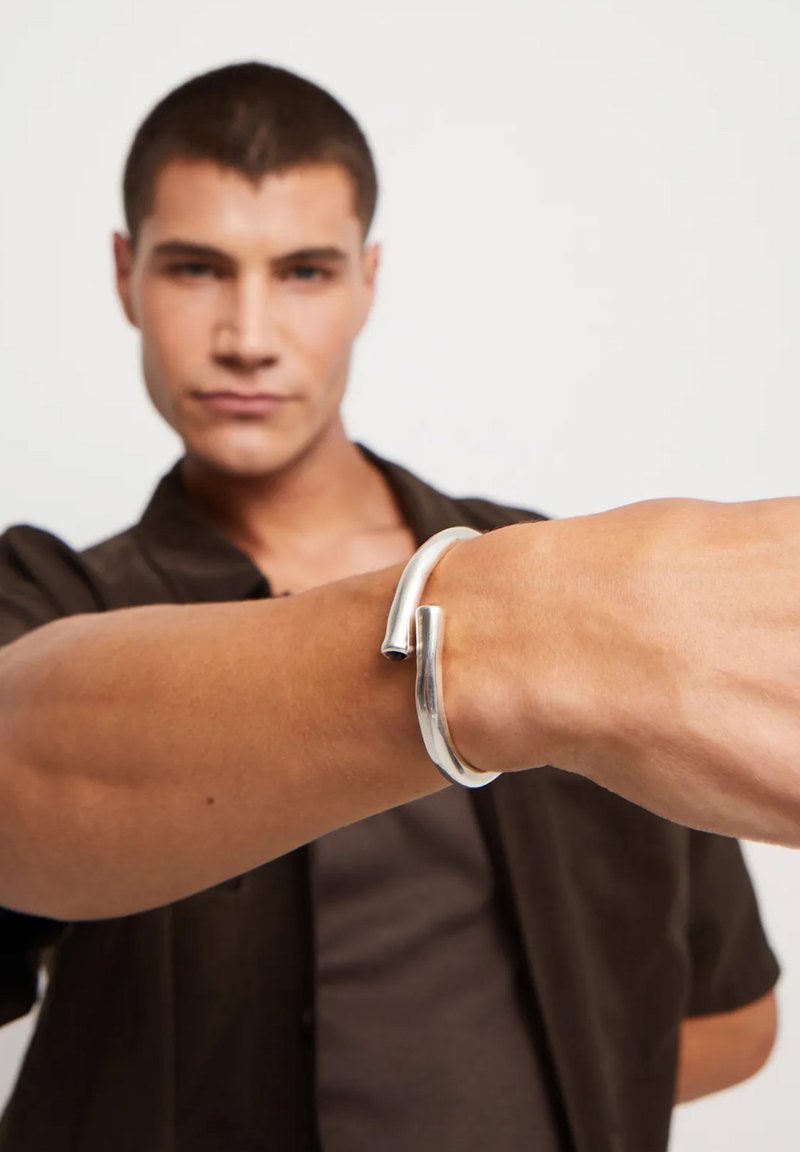 Bracelet en argent avec une finition lisse et polie, arborant un design incurvé et un détail d'ouverture distinctif. Porté sur un poignet masculin.