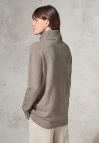 Hellgrauer Rollkragenpullover mit lockerer Passform, weichem Material, geripptem Kragen und Saum, kombiniert mit hellen Hosen.