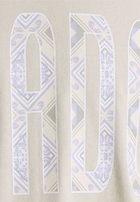 Ljusbeige tyg har stora stiliserade bokstäver med ett mönstrat överlägg i mjuka lavendel- och gröntoner, mot en texturerad bakgrund.