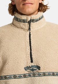 Beige fleece sweater med en halv-lynlås krave. Har broderet logo og dekorativt tribal-mønstret pynt. Blødt, tekstureret stof.