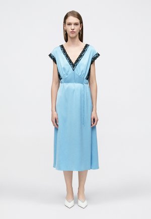 Kobieta stojąca w jasnoniebieskiej sukience midi z czarną koronką przy dekolcie w kształcie litery V i na rękawach, w połączeniu z białymi, spiczastymi butami.