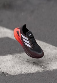 Zwarte sport schoen met een bovenwerk van mesh, drie witte strepen en een rode tussenzool. Geprofileerd oppervlak met een gestroomlijnd ontwerp.