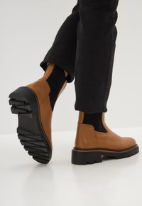 Botas de cuero marrón hasta el tobillo con paneles laterales elásticos, suela de goma negra texturizada y tacón grueso. Combinadas con pantalones negros ajustados.