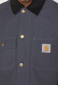 Giacca da lavoro grigia con colletto in velluto a coste nero, due tasche frontali, bottoni metallici e una toppa con logo cucita, caratterizzata da un design circolare.