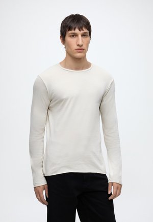 Joven con el cabello corto y oscuro, vistiendo una camisa blanca ajustada de manga larga y pantalones negros, de pie frente a un fondo claro y liso.