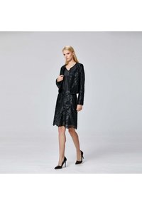 Femme blonde portant une jupe en dentelle florale noire et une veste assortie avec un top noir et des talons hauts noirs sur un fond clair uniforme.