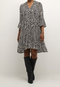 Šaty s leopardím potiskem v černobílé barvě, s výstřihem do V, tříčtvrtečními zvonovými rukávy a rozšířeným střihem, v kombinaci s černými kozačkami po kolena.