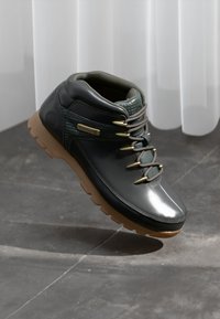 Botină sport de culoare gri închis cu finisaj lucios, talpă din cauciuc și accente verzi. Prezentă găuri pentru șireturi din metal și o guler căptușit. Logo-ul Timberland este vizibil.