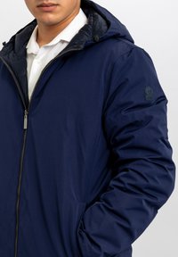 Giacca imbottita blu navy con chiusura lampo e cappuccio; presenta un esterno liscio, polsini elasticizzati e un logo sul braccio sinistro.