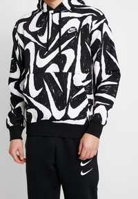 Svart och vit Nike hoodie med ett virvlande abstrakt mönster, snörning i huvan och känguruficka; tillverkad av mjukt material med ribbade ärmslut.
