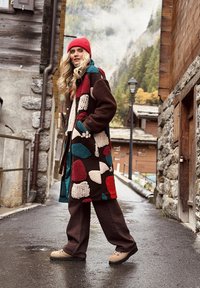 Žena v farebnom patchworkovom kabáte a červenej čiapke kráča po mokrej ulici v horskej dedine so skalnatými a drevenými budovami.