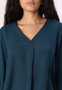 Teal lichtgewicht stoffen top met een V-halsontwerp, met een subtiele textuurafwerking en een losse pasvorm, die tot halverwege de borst reikt.