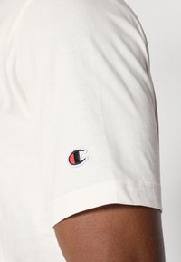 Weißes Baumwoll-T-Shirt mit kurzem Arm. Auffälliger Logo-Patch in Rot und Schwarz am linken Ärmel. Glatte Textur mit klassischer Passform.