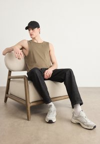 Brun uten ermer skjorte, svart caps, svarte jeans, og grå sneakers. Sitter på en beige sofa med buet armlene og trechassis.