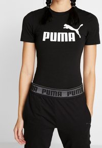 Młoda osoba z plecionymi włosami, ubrana w czarny t-shirt Puma i spodnie z markowym elastycznym pasem, stojąca z jedną ręką w kieszeni.