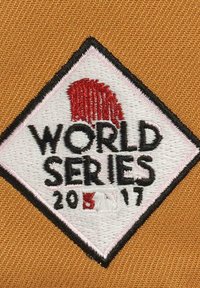 Embroidered patch featuring a diamond shape with "WORLD SERIES 2017" text, red accent, and white background on a textured mustard fabric.