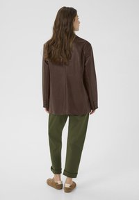 Frau mit langen Haaren, die eine braune Lederjacke, grüne hochgekrempelte Hosen und beigen Sneakers trägt, steht mit dem Rücken zu einem schlichten Hintergrund.